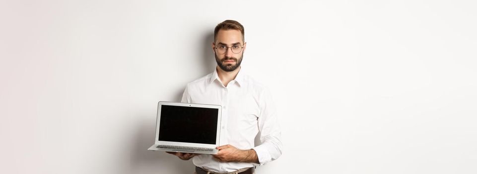 Confident manager demonstrating presentation on screen, showing laptop display and looking serious, standing over white background.