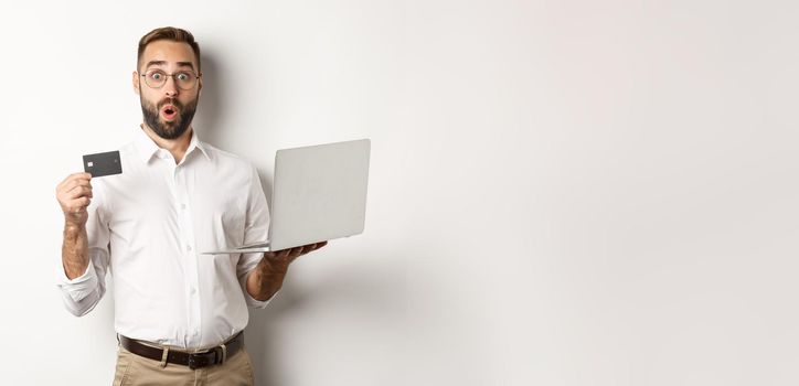 Online shopping. Surprised man holding laptop and credit card, shop internet store, standing over white background.