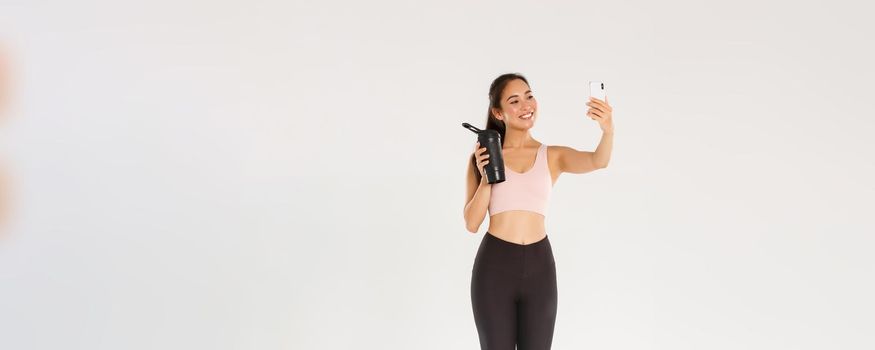 Full length of smiling slim and healthy asian girl taking selfie fitness training session, female blogger and athelte showing water bottle on camera of mobile phone, standing over white background.