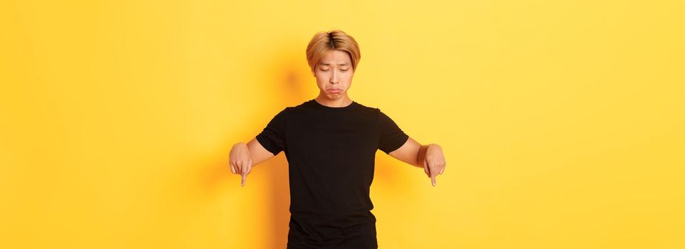 Portrait of gloomy and upset asian man sulking with regret, pointing and looking down at banner, yellow background.