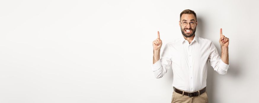 Doubtful and worried man looking and pointing fingers up, grimacing hesitant, standing over white background.