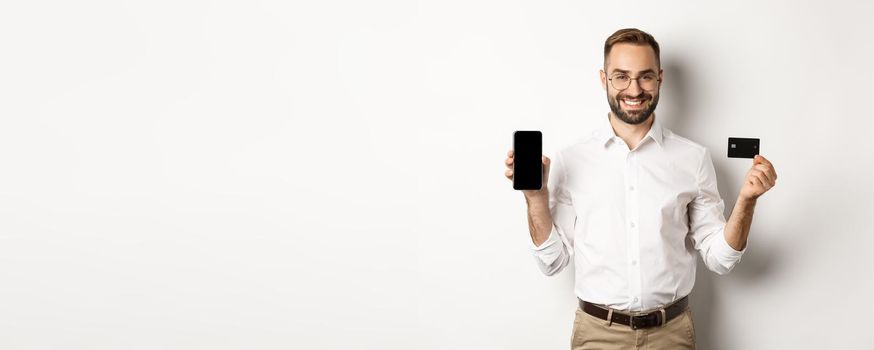 Business and online payment. Smiling handsome man showing mobile screen and credit card, standing over white background.