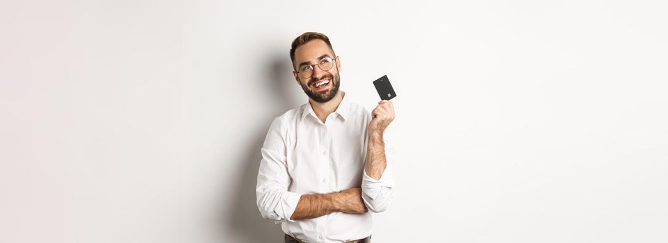 Image of handsome man thinking about shopping and holding credit card, looking at upper left corner thoughtful, white background.