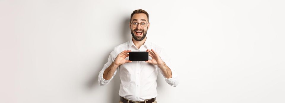 Excited handsome man showing mobile phone screen, looking amazed, standing over white background.