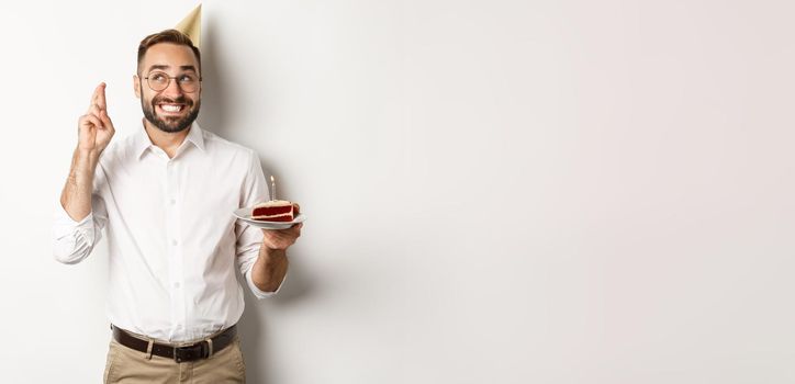 Holidays and celebration. Happy man having birthday party, making wish on b-day cake and cross fingers for good luck, standing against white background.