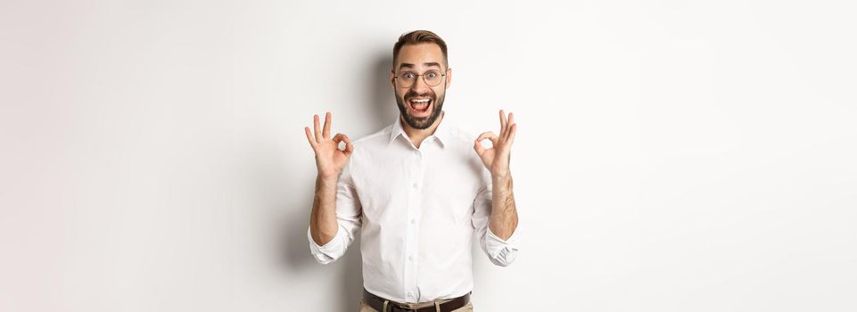 Amazed entrepreneur showing okay sign and looking happy, satisfied with product, standing over white background. Copy space