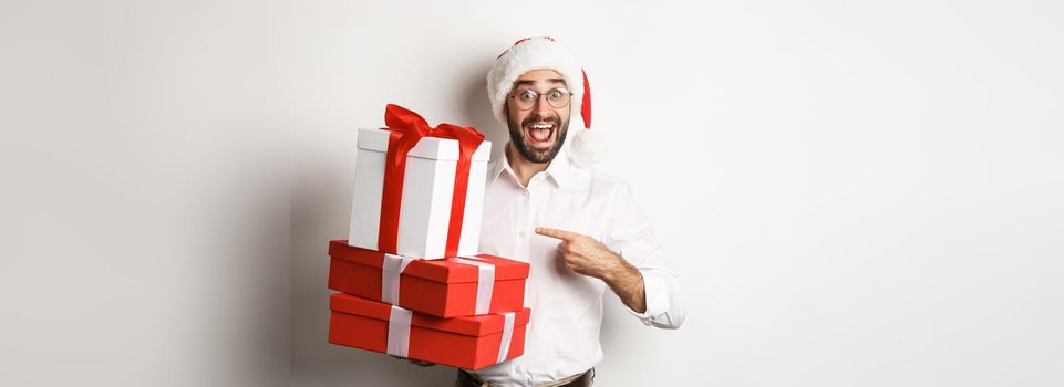 Merry christmas, holidays concept. Surprised man receive xmas gifts, pointing at presents and smiling happy, wearing santa hat, white background.