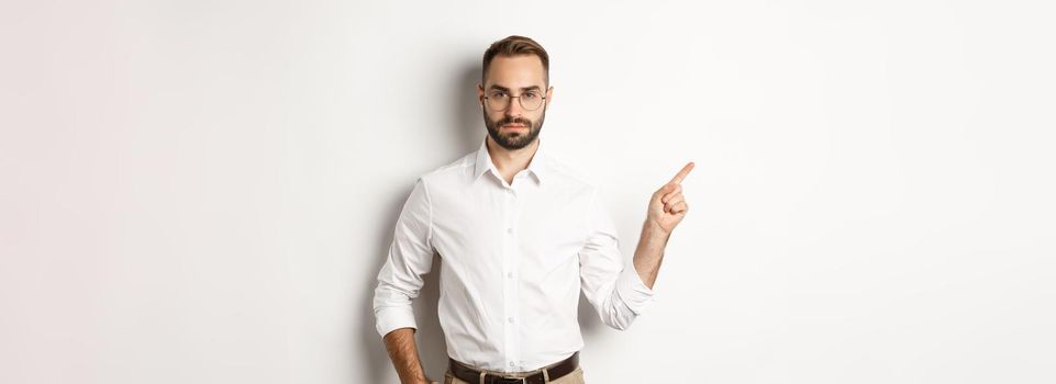 Serious bearded man pointing finger left, showing advertisement, standing over white background.