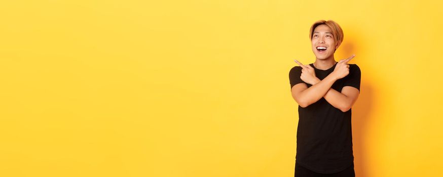 Portrait of happy smiling asian man with blond hair, looking amused left, pointing sideways at two variants, yellow background.