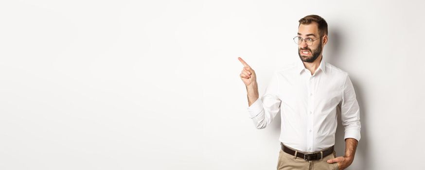 Young bearded guy seeing something disturbing, cringe while pointing finger left at promo offer, standing awkward against white background.