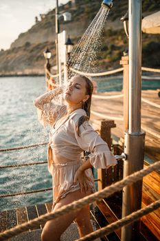 Beautiful young woman with a perfect figure in the shower in the rays of the sunset by the sea. The girl with a golden tan on vacation. Selective focus