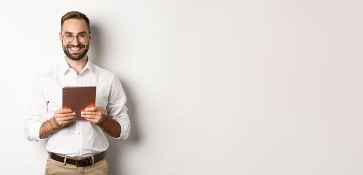 Handsome bearded man in glasses using digital tablet, smiling satisfied, standing against white background.