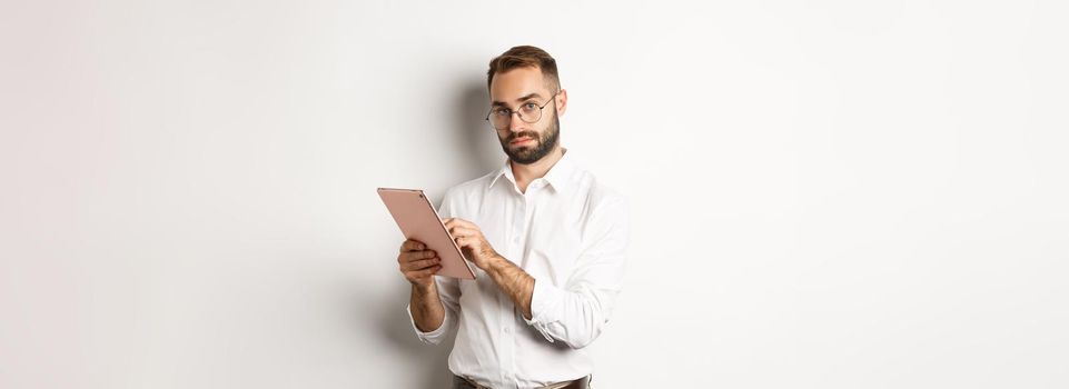 Handsome businessman doing job on digital tablet, reading something, standing over white background.