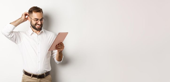 Confused male manager looking puzzled at digital tablet, scratching head doubtful, standing over white background.