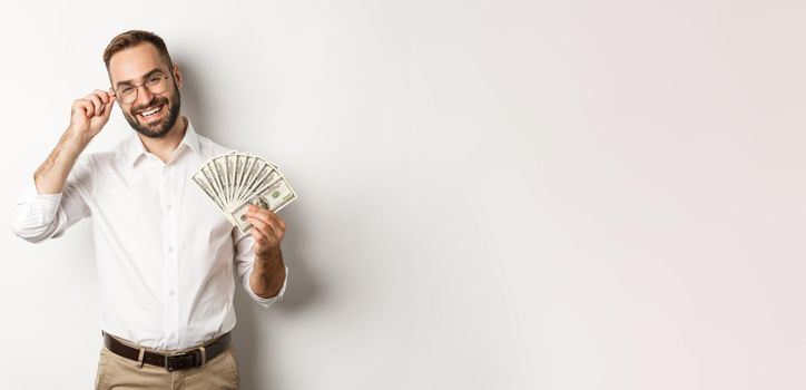 Handsome successful businessman holding money, fixing glasses on nose, standing over white background.