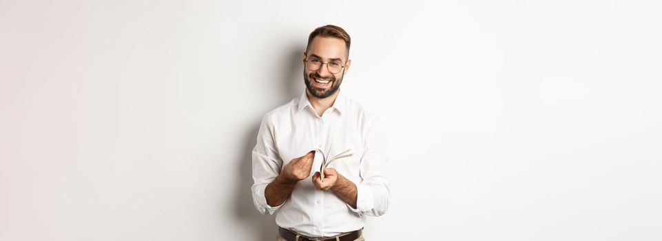 Successful businessman in white shirt, counting money and smiling satisfied, standing over white background.