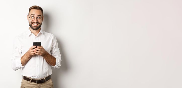 Thouthful handsome manager using mobile phone and thinking about answering message, looking at upper left corner and smiling, standing over white background.