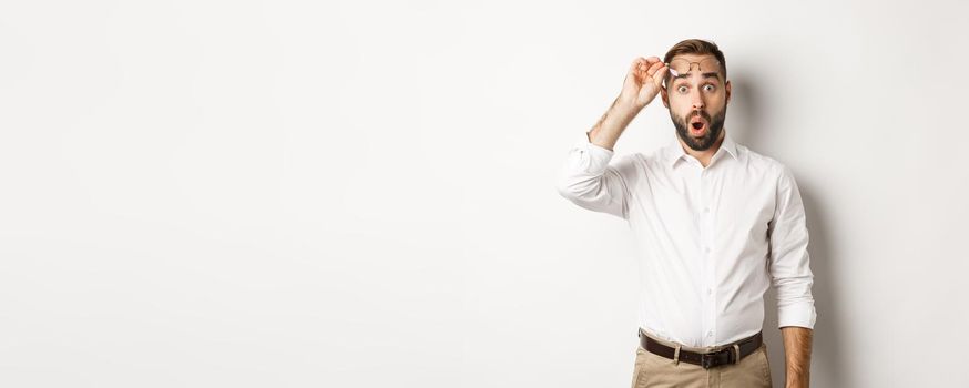 Surprised businessman taking-off his glasses, looking with amazement at camera, standing over white background.