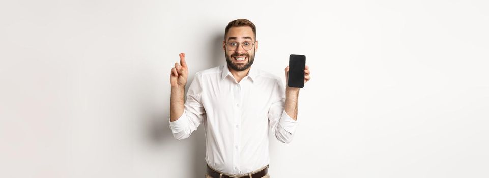 Hopeful young business man showing mobile screen, holding fingers crossed, waiting for online results, standing over white background.