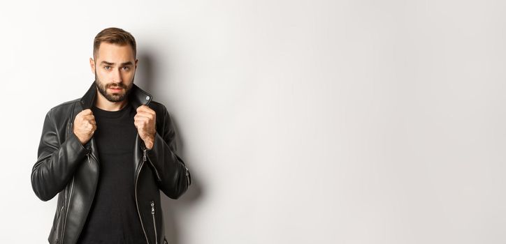 Image of handsome and confident man putting on leather biker jacket, standing against white background.
