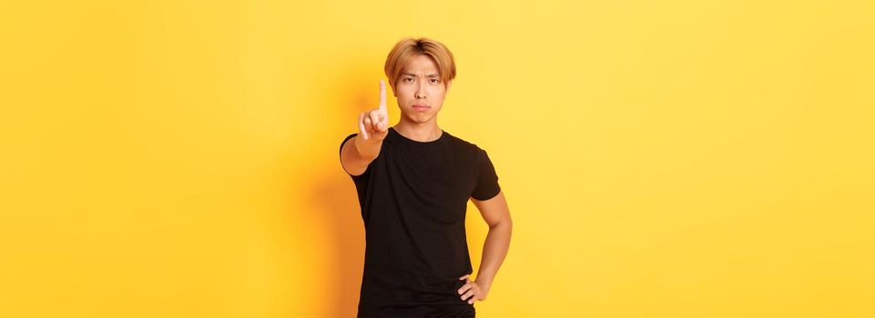 Portrait of serious-looking disappointed asian man shaking finger to scold someone, standing yellow background.