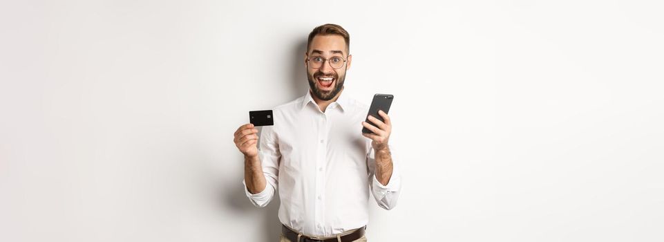 Business and online payment. Excited man paying with mobile phone and credit card, smiling amazed, standing over white background.