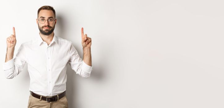 Young bearded businessman pointing fingers up, showing promo offer, standing over white background. Copy space