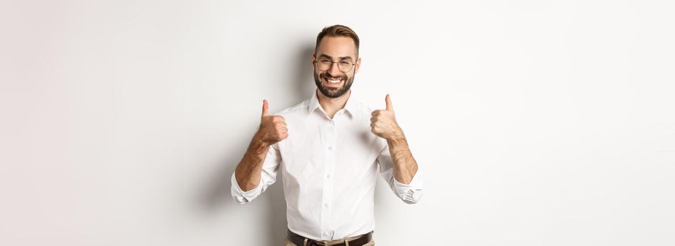 Successful businessman praising good work, showing thumbs up and smiling satisfied, standing over white background.