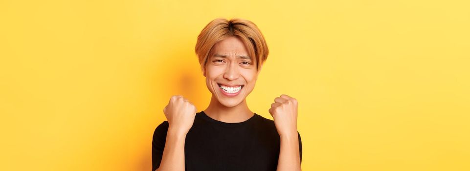 Close-up of lucky happy asian guy winning prize, making fist pump gesture and smiling satisfied, triumphing over achievement, standing yellow background.
