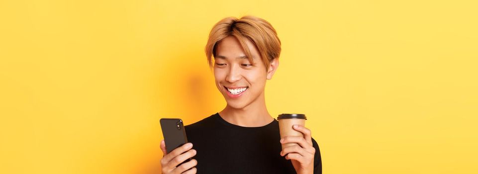 Close-up of handsome stylish asian male student, using mobile phone and drinking coffee. Guy messaging on smartphone, standing over yellow background.