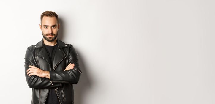 Handsome man wearing leather jacket, looking confident at camera, standing over white background.
