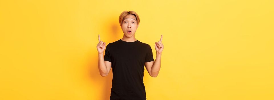 Portrait of astonished asian guy in black t-shirt, open mouth fascinated, pointing fingers up, yellow background.