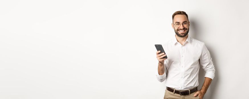 Handsome manager using smartphone and smiling pleased, sending text message, standing over white background.