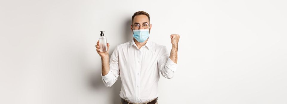 Covid-19, social distancing and quarantine concept. Cheerful manager in medical mask showing hand sanitizer, standing over white background.