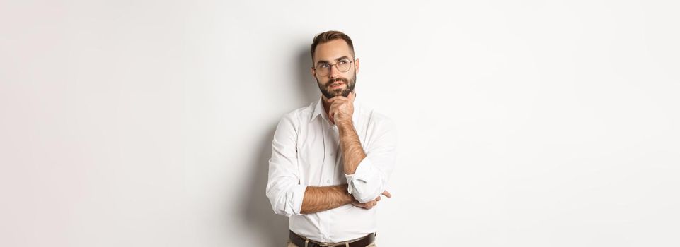 Thoughtful businessman in glasses making plan, looking at upper left corner and thinking, standing against white background.