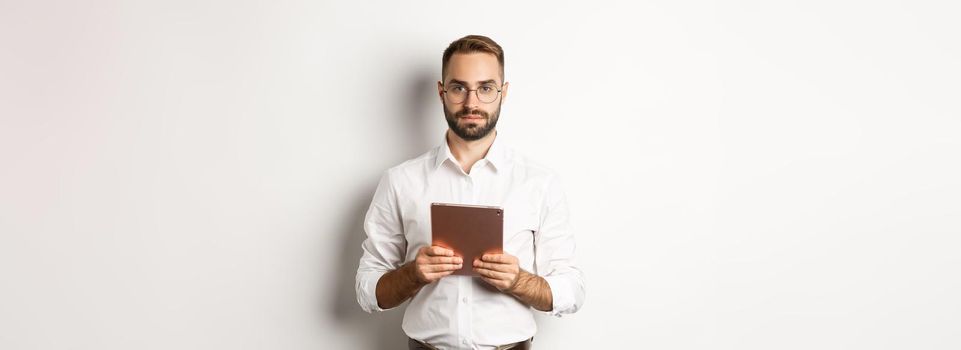 Serious employer working with digital tablet, reading in glasses, standing over white background.