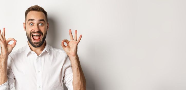 Close-up of amazed caucasian guy agreeing, showing okay signs and looking impressed, recommending product, white background.