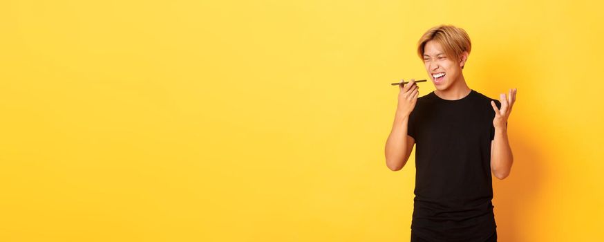 Portrait of angry asian man yelling mad at mobile phone speaker, record furious voice message, standing yellow background.