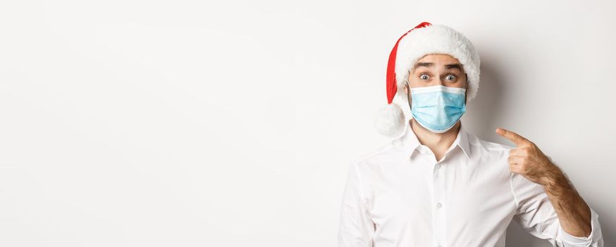 Concept of coronavirus, quarantine and winter holidays. Excited guy in santa hat pointing at his face mask, standing over white background.
