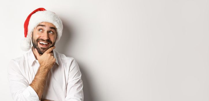 Party, winter holidays and celebration concept. Close-up of happy man planning christmas gift list, wearing santa hat, looking at upper left corner thoughtful, white background.