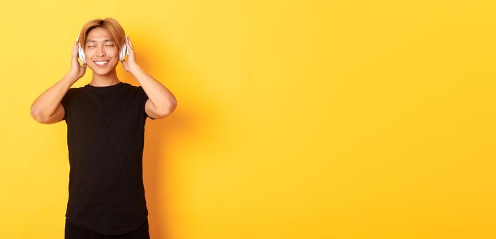 Relaxed and pleased handsome asian guy close eyes from satisfaction while listening music in wireless headphones, standing yellow background.