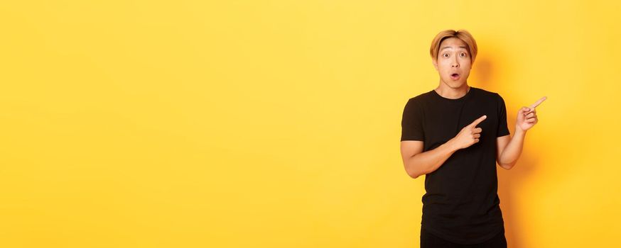 Portrait of curious and amused blond asian guy, pointing fingers upper right corner, showing way, standing yellow background.