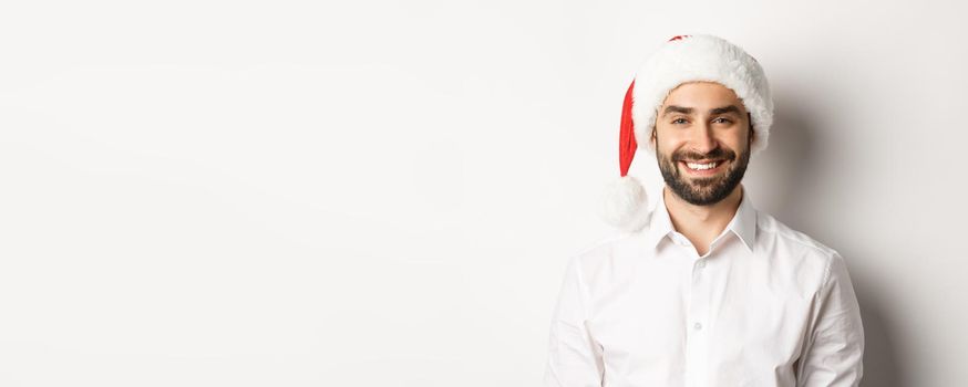 Close-up of happy bearded man celebrating christmas, wearing santa party hat and smiling, standing over white background.