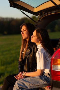 Mother and daughter camping on a hill and admiring the sunset while sitting in the car trunk