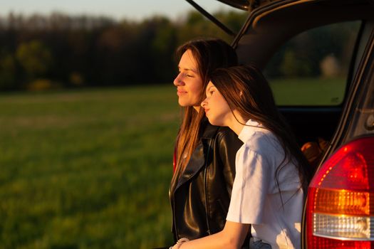 Mother and daughter camping on a hill and admiring the sunset while sitting in the car trunk