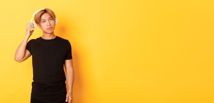 Young handsome asian guy with blond hair, take-off headphones to listen you, standing yellow background.