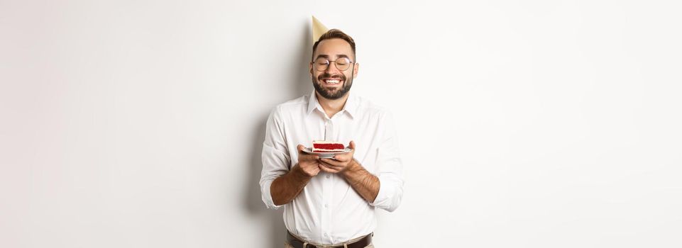 Holidays and celebration. Happy man having birthday party, making wish on b-day cake and smiling, standing against white background.