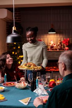 Family gathering at christmas festive dinner table, winter holiday celebration, african american woman holding traditional chicken dish. Happy diverse people celebrating xmas at home party