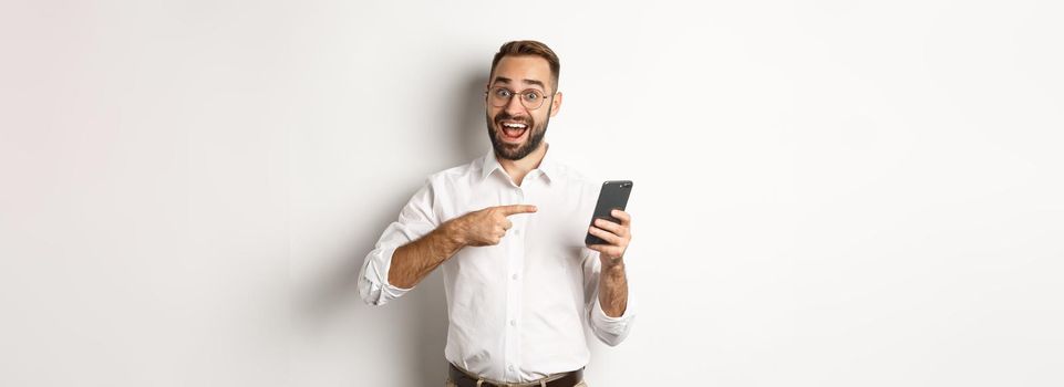 Man looking excited and pointing finger at mobile phone, showing good online offer, standing over white background.