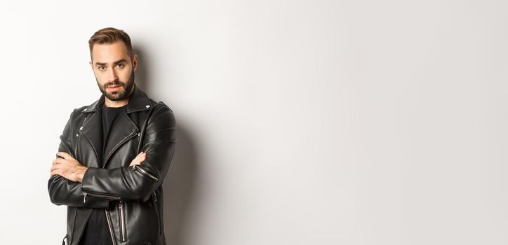 Attractive macho man with beard, wearing leather jacket smiling, looking confident with hands crossed on chest, white background.